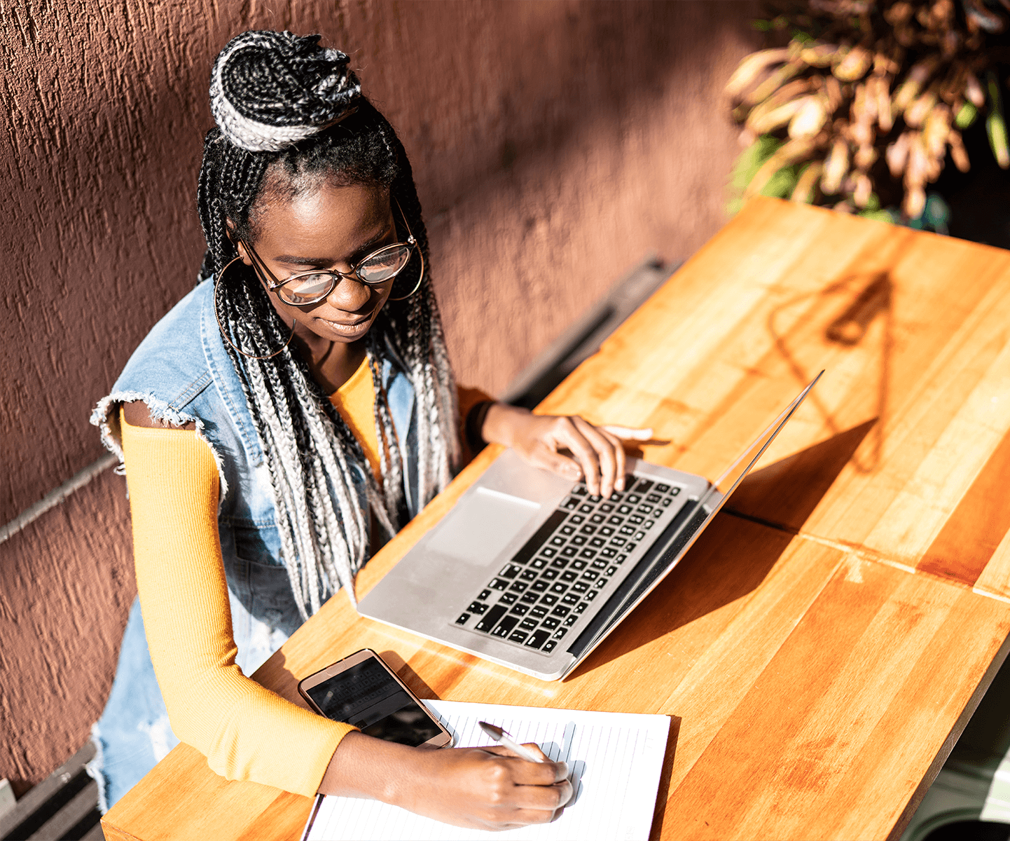 African American woman at a table with her laptop taking notes on a note pad.